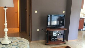 Un salon avec un téléviseur posé sur un meuble, une table recouverte d’une nappe à motifs floraux et un banc en bois.