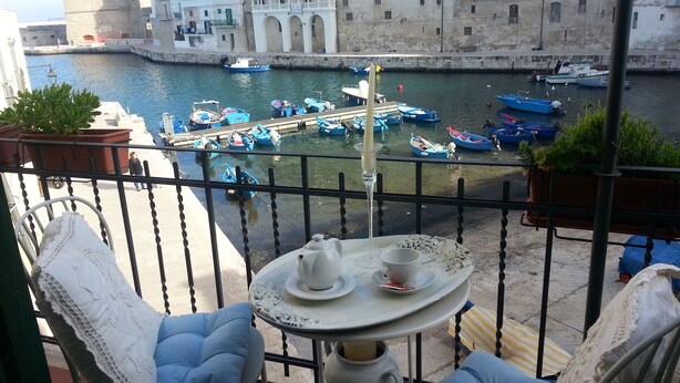A balcony with a table set for two, overlooking a harbor with boats and historic buildings.