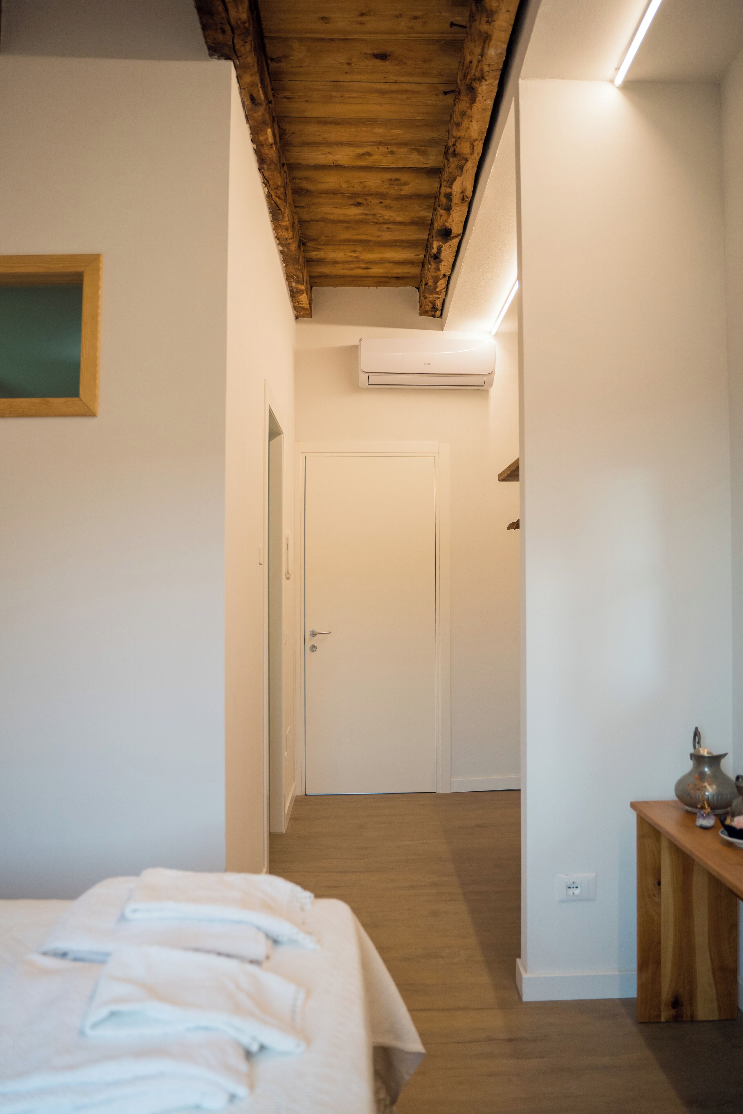 A bedroom with a bed, a wooden bench, an air conditioning unit, and a door leading to another room.
