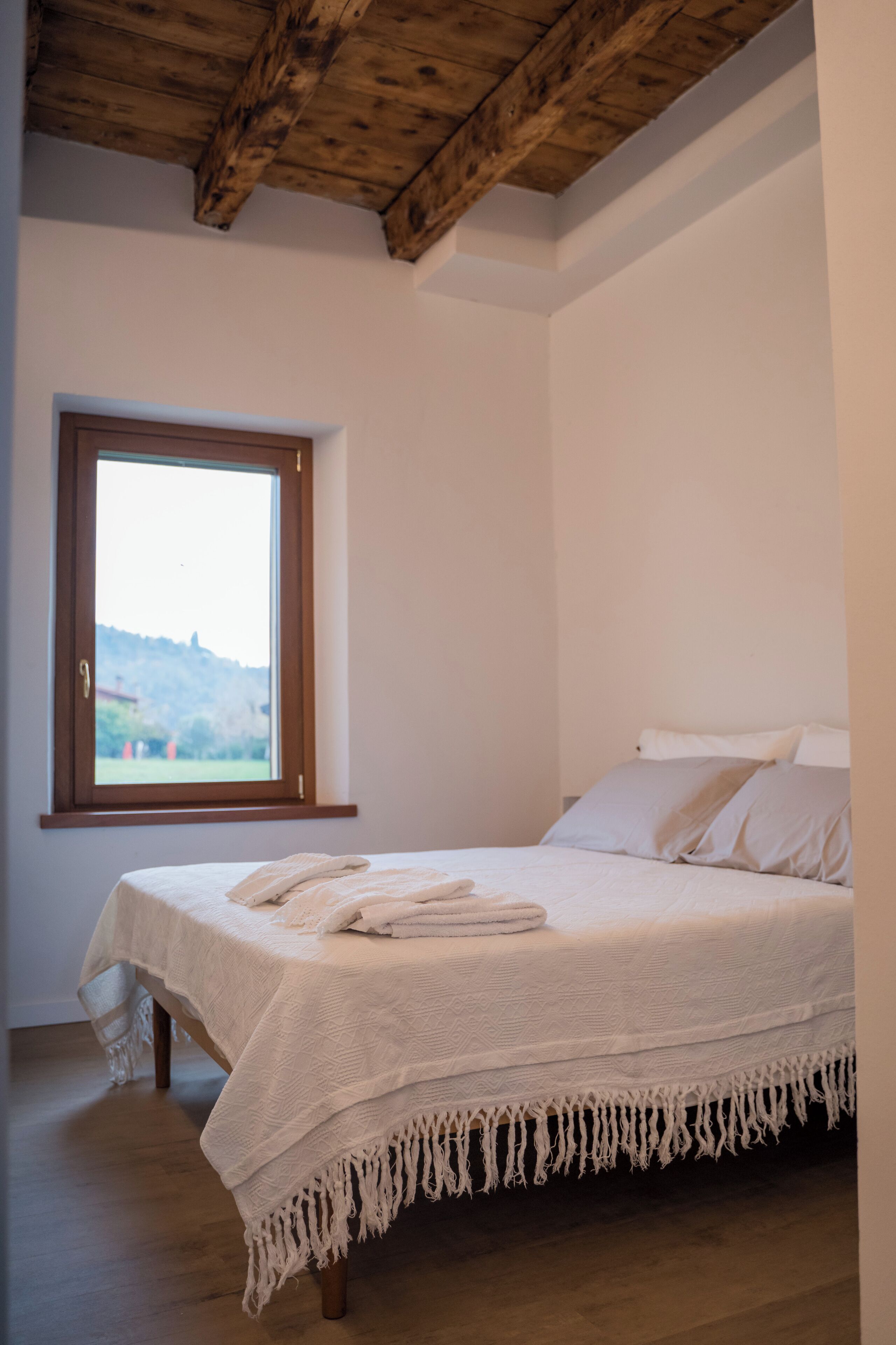 A bedroom with a large window, a bed with white linens, wooden beams on the ceiling, and a view of a landscape outside.