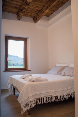 Ein Schlafzimmer mit großem Fenster, einem Bett mit weißen Leinen, Holzbalken an der Decke und Blick auf eine Landschaft.