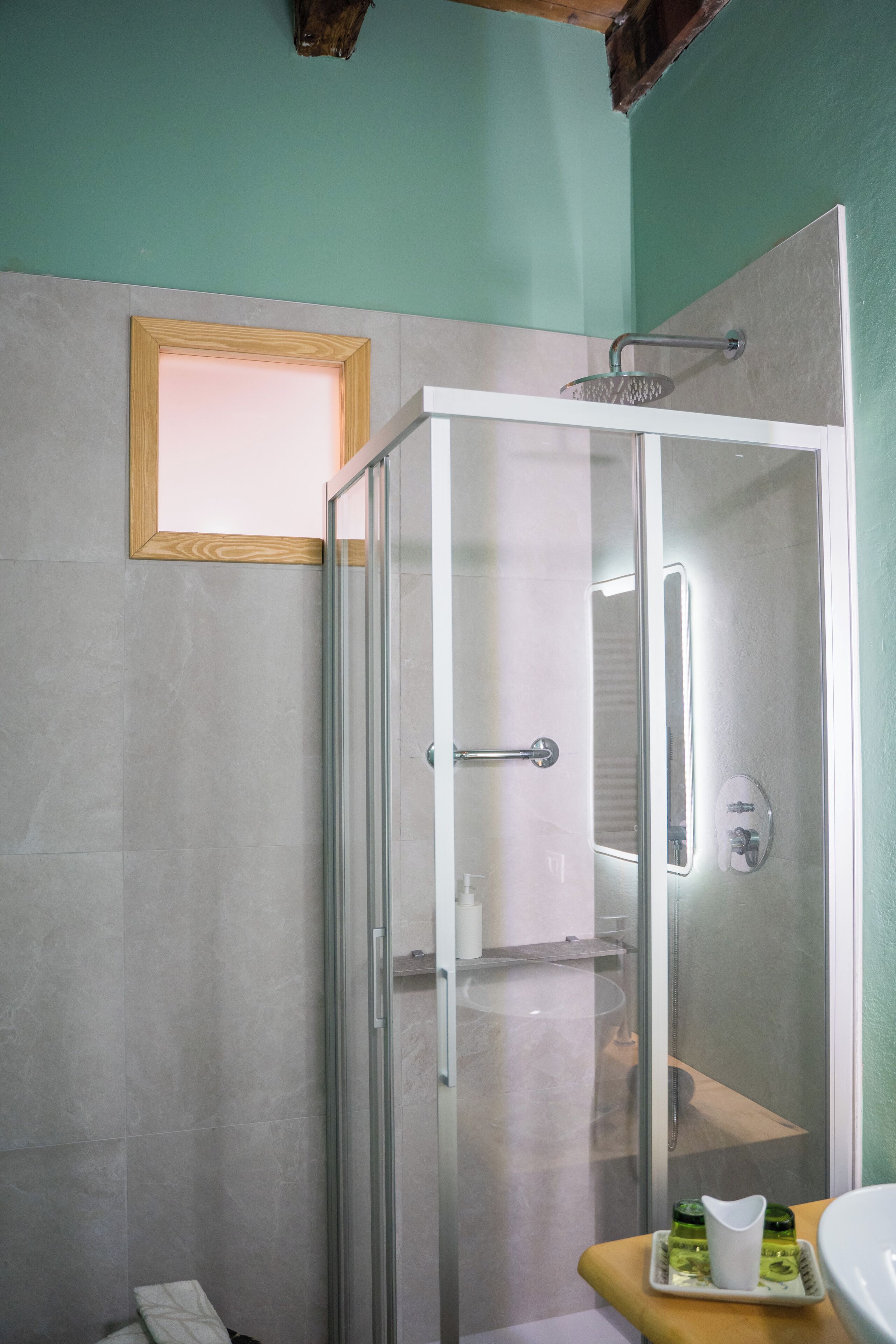 Doccia vetrata con soffione a mano, lavandino bianco con specchio e ripiano in legno con articoli da toilette.