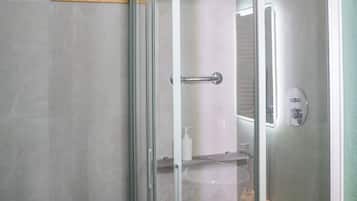 Doccia vetrata con soffione a mano, lavandino bianco con specchio e ripiano in legno con articoli da toilette.