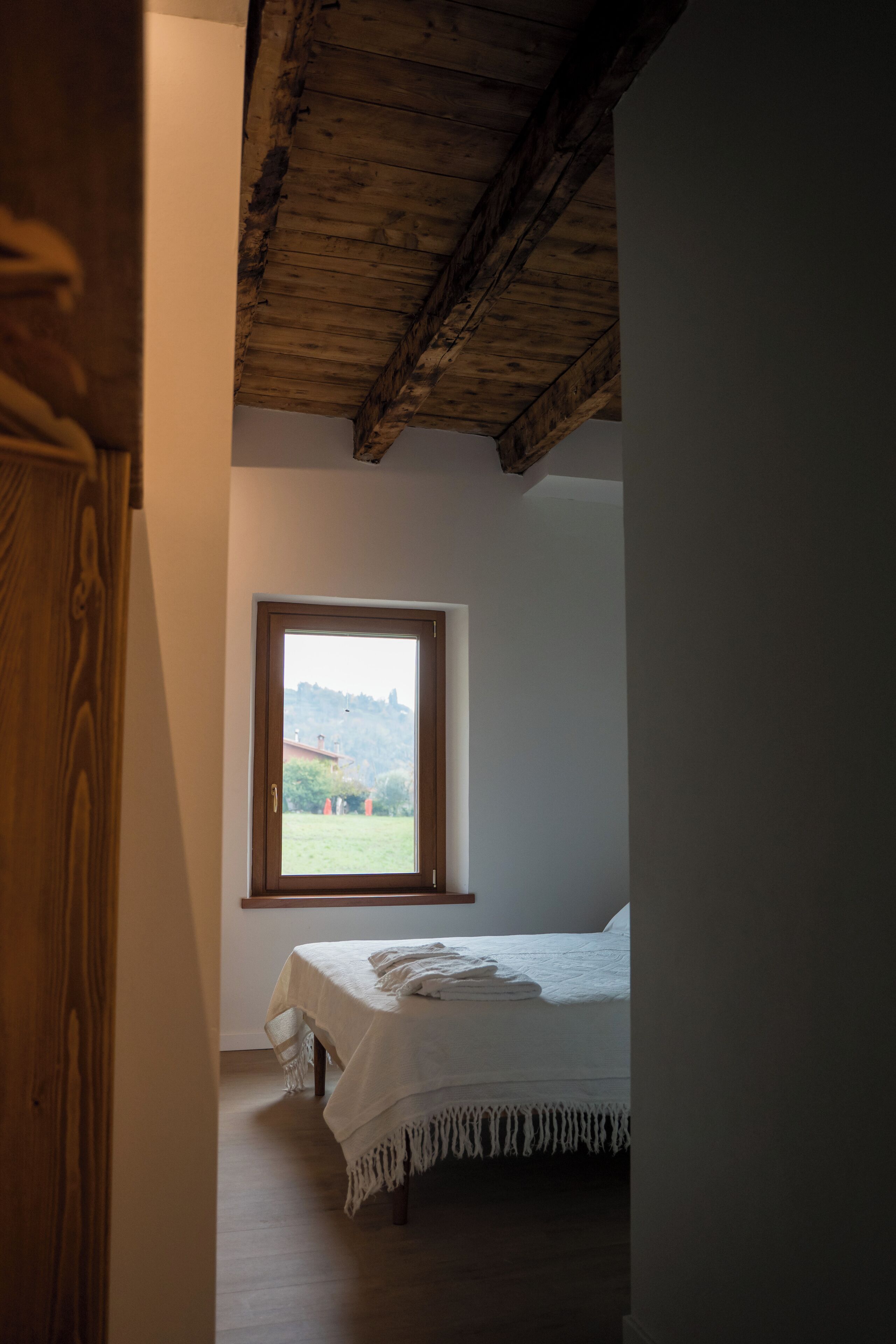 Un dormitorio con techo de madera, una ventana con vista a una colina y una cama con ropa blanca.