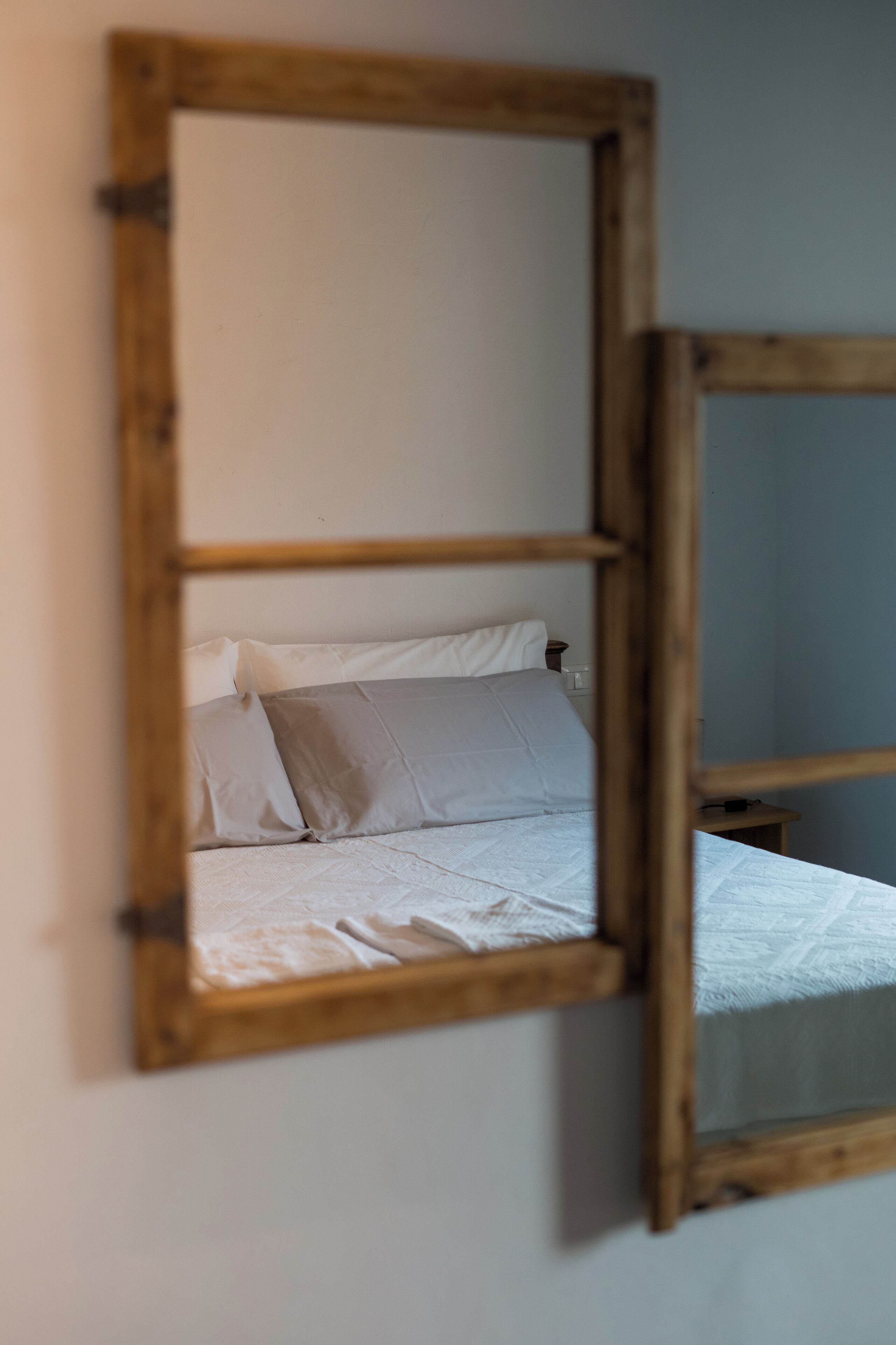 Een slaapkamer met een spiegel met houten lijst, die een bed met witte lakens en een grijs kussen weerspiegelt.
