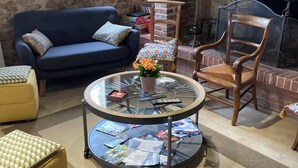 Ein gemütliches Wohnzimmer mit Kamin aus Backstein, einem blauen Sofa, einem Holzstuhl, einem Glastisch mit Zeitschriften und einem gelben Hocker.