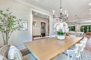 A spacious dining area with a long wooden table, white chairs, a chandelier, and a potted orchid. - 19 Sandcastle, Stunning New House, Free Heated Pool, 3 Blocks to Beach, 3D Home Theater, Elevator (Hilton Head Island)