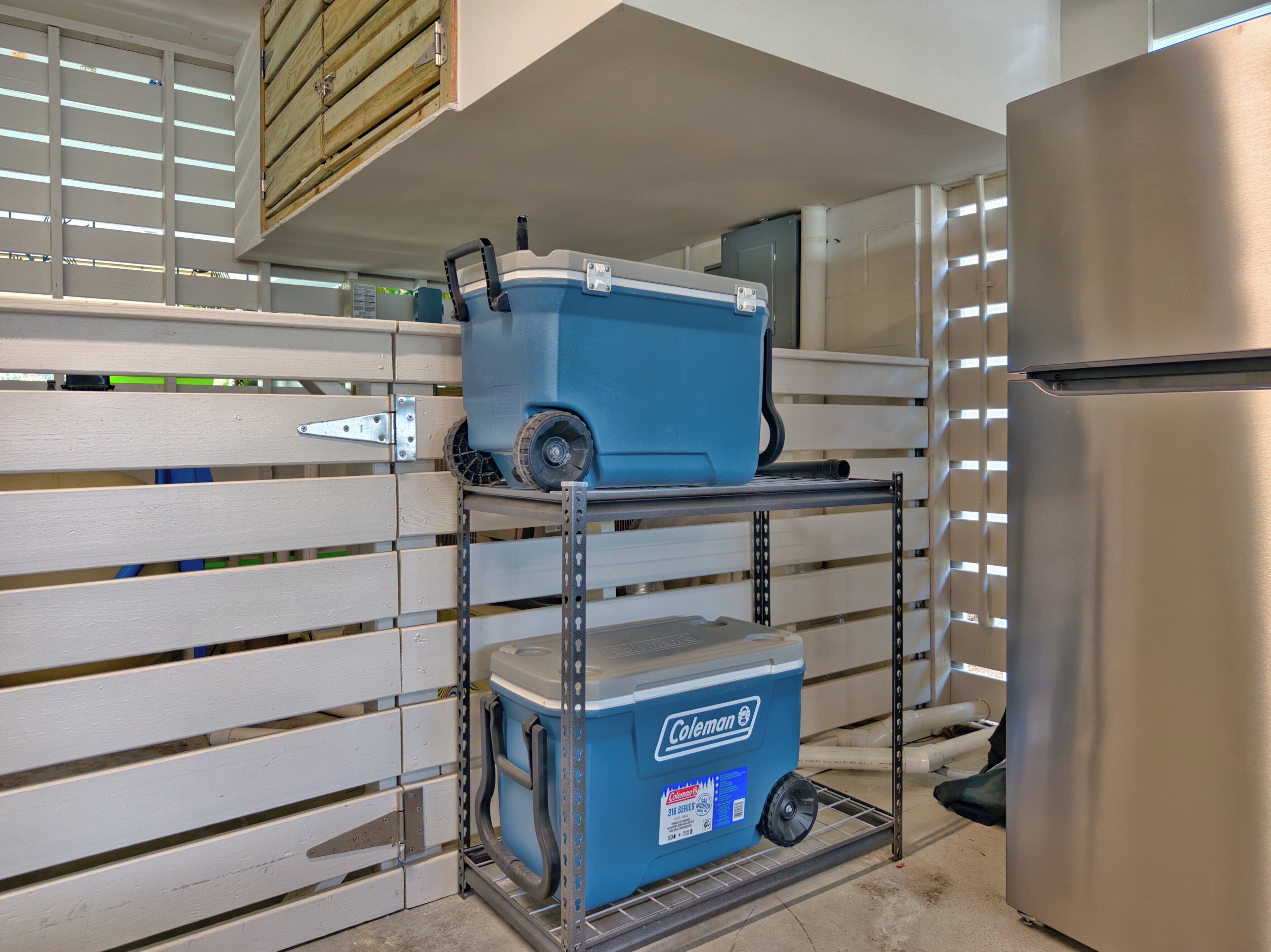 Una cuina amb un refrigerador d'acer inoxidable, una nevera Coleman blava sobre una prestatgeria de metall i un armari de metall amb una porta de fusta.