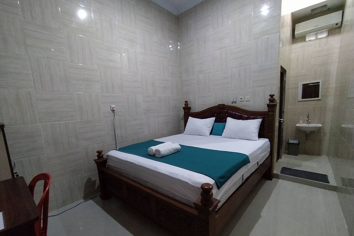 A neatly made bed with a teal bedspread and white linens, a wooden headboard, and a small bedside table with a red chair.