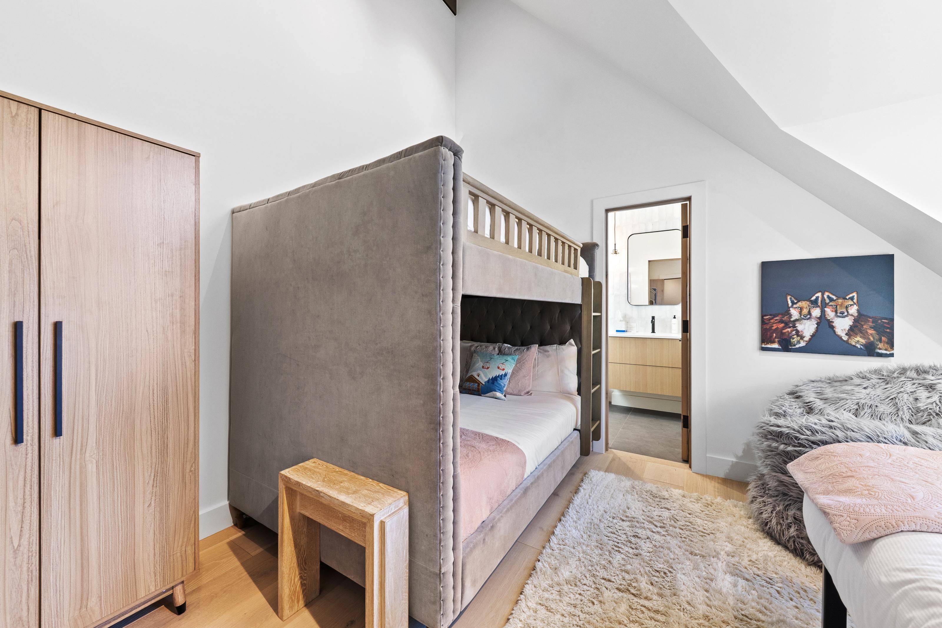 A bedroom with a bunk bed, a wooden wardrobe, a small wooden stool, a fluffy rug, and a painting on the wall.