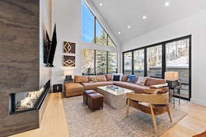 Un salon moderne doté d’une cheminée, d’un grand canapé d’angle, d’une table basse et offrant une vue sur un paysage enneigé grâce à de grandes fenêtres.