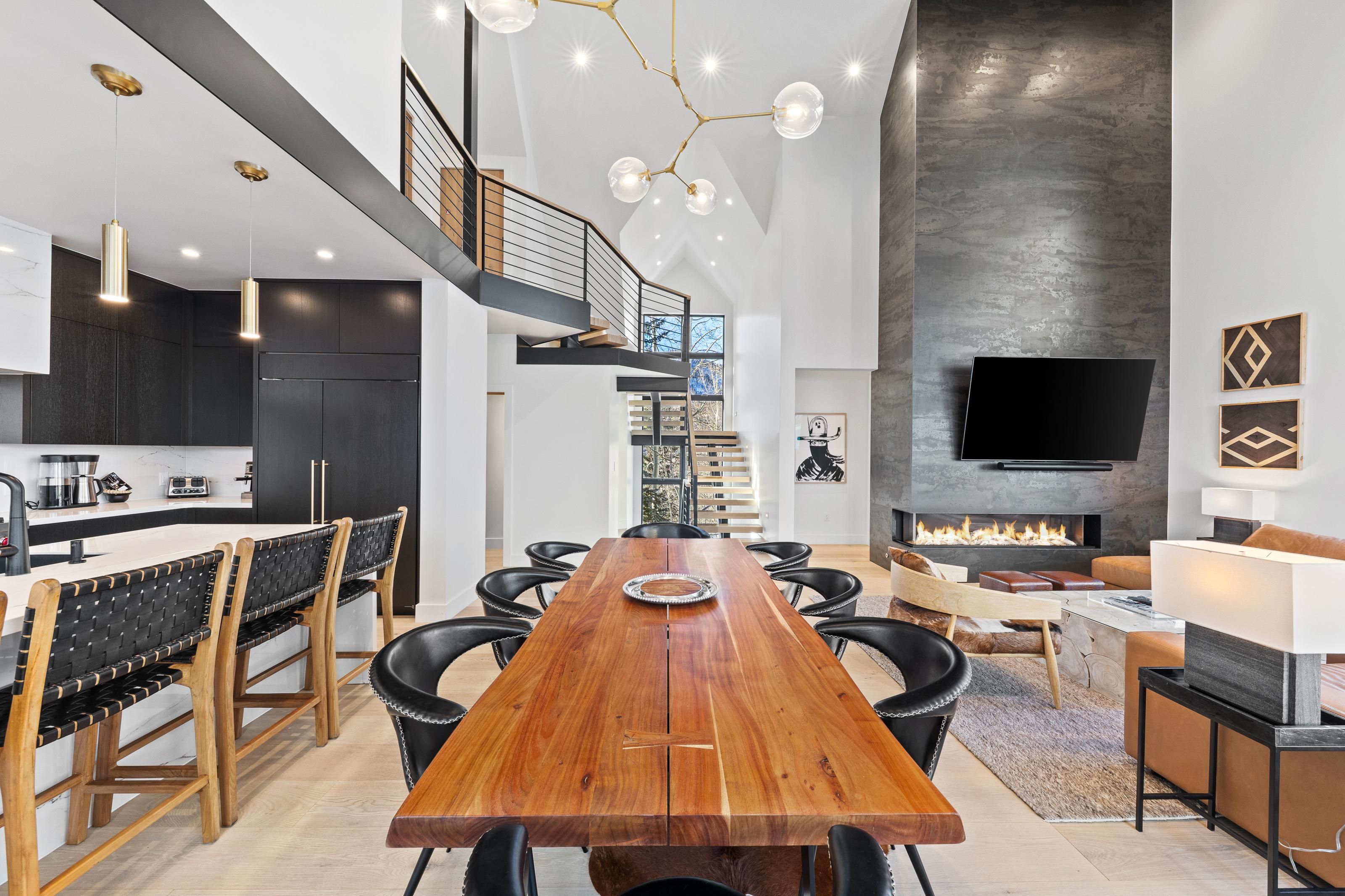 Un intérieur moderne avec une table à manger en bois, des chaises en cuir noir, une cuisine aux meubles sombres, un escalier et une cheminée.