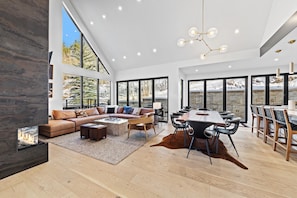 Un vaste espace de vie doté d’une cheminée, d’un grand canapé, d’une table à manger avec des chaises et de grandes fenêtres offrant une vue sur un paysage enneigé.