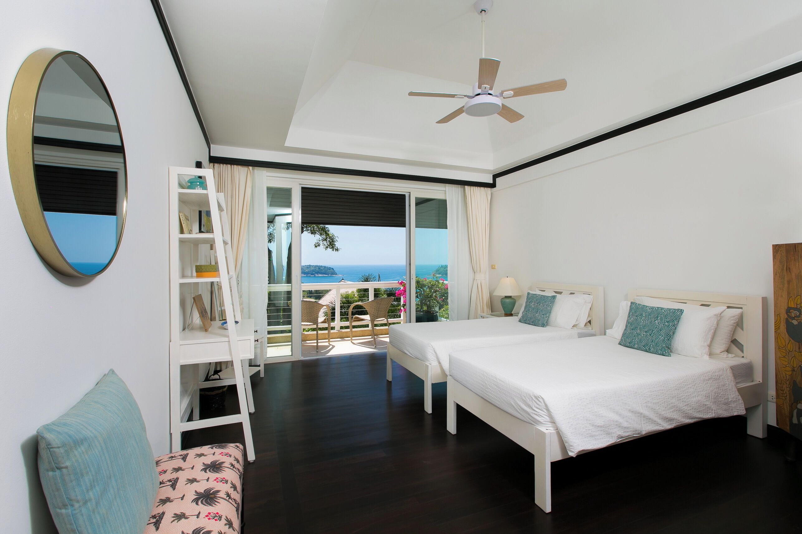 A bedroom with a bed, a desk, a chair, a ceiling fan, and a view of the sea.