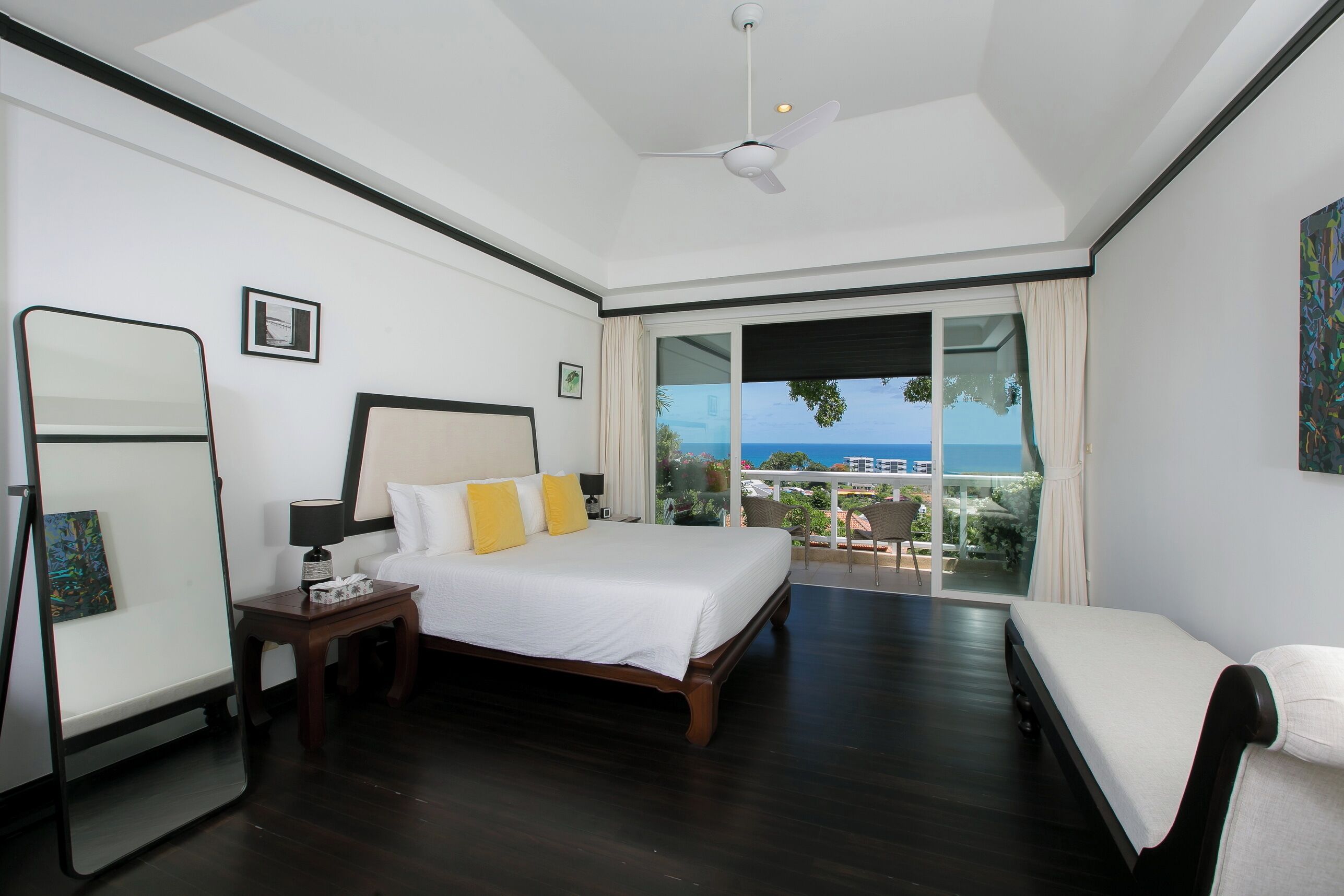 A hotel room with a bed, a bedside table, a mirror, a window with a view, and a ceiling fan.