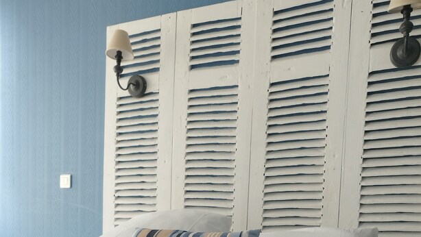 Una cama bien hecha con toallas blancas enrolladas, un cojín a rayas y ropa de cama blanca, frente a una pared azul con cabecera de madera.