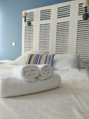 A neatly made bed with rolled white towels, a striped pillow, and white bedding against a blue wall with a wooden headboard. - Hôtel les Thermes (Jonzac)