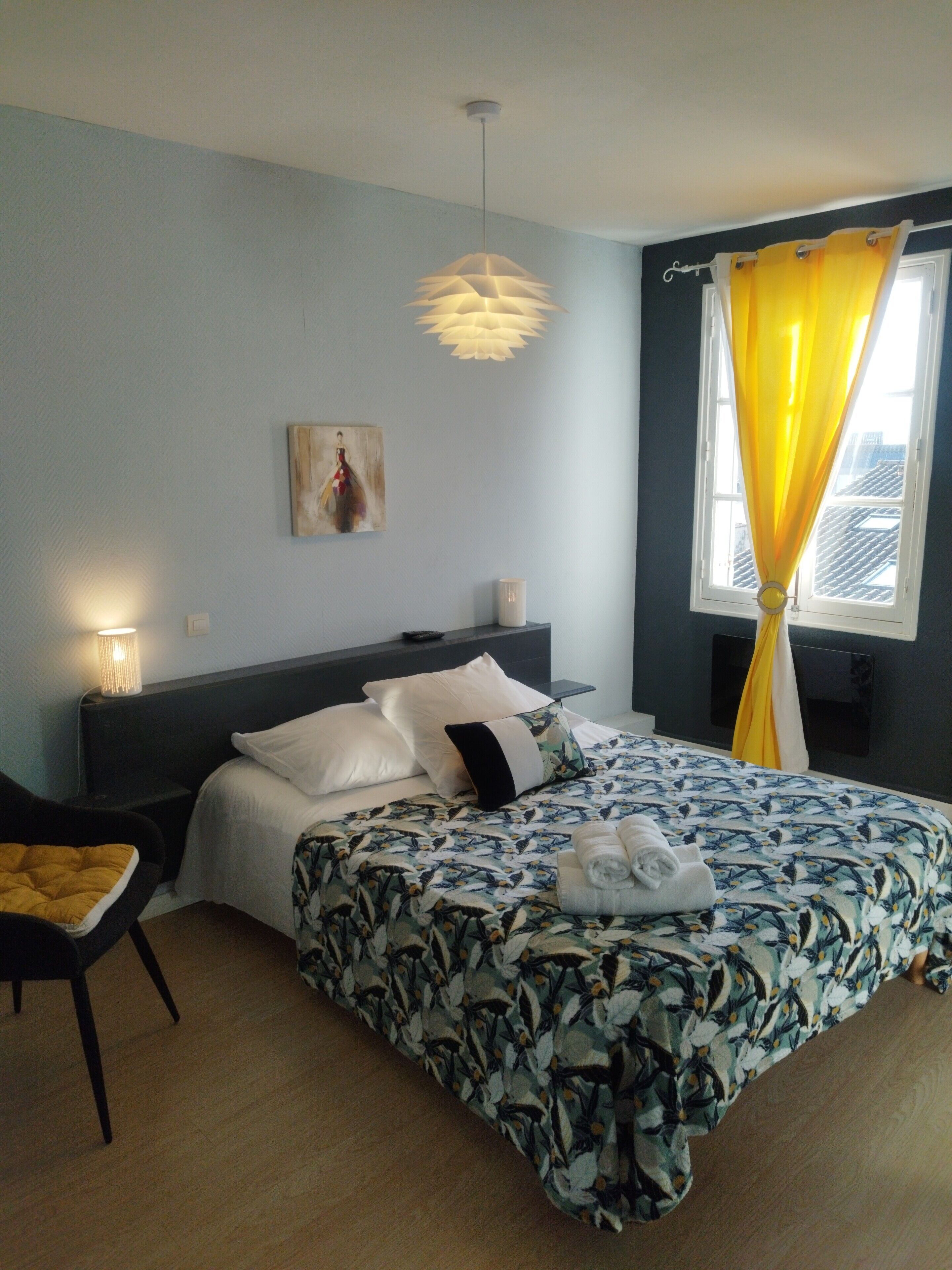 Un dormitorio con cama, una mesita de noche, una silla, un cuadro en la pared y una ventana con cortinas.