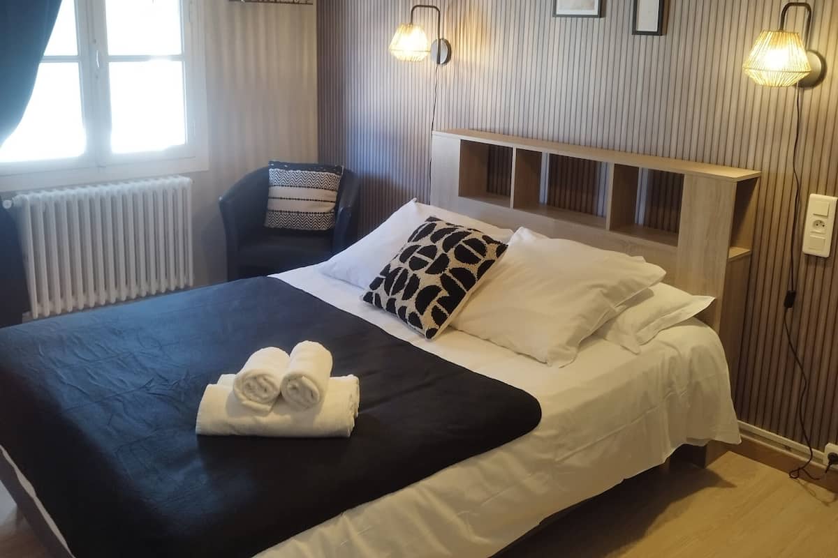 Cama bem arrumada com colcha preta e branca, um par de toalhas enroladas e cabeceira com prateleiras embutidas. Há duas luminárias fixadas na parede e uma janela com cortinas.