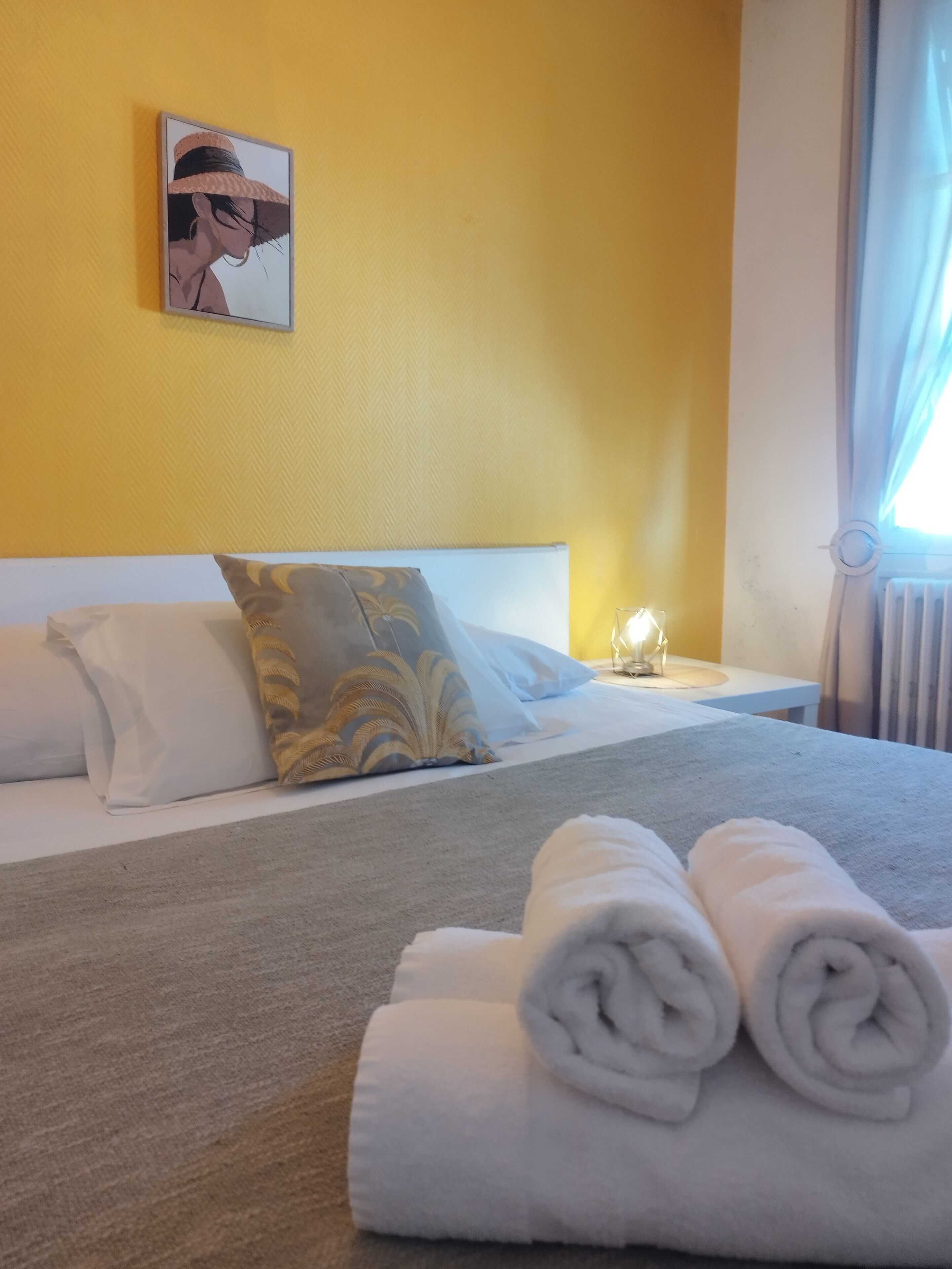 A hotel room with a bed, pillows, a nightstand with a lamp, and a window with curtains.