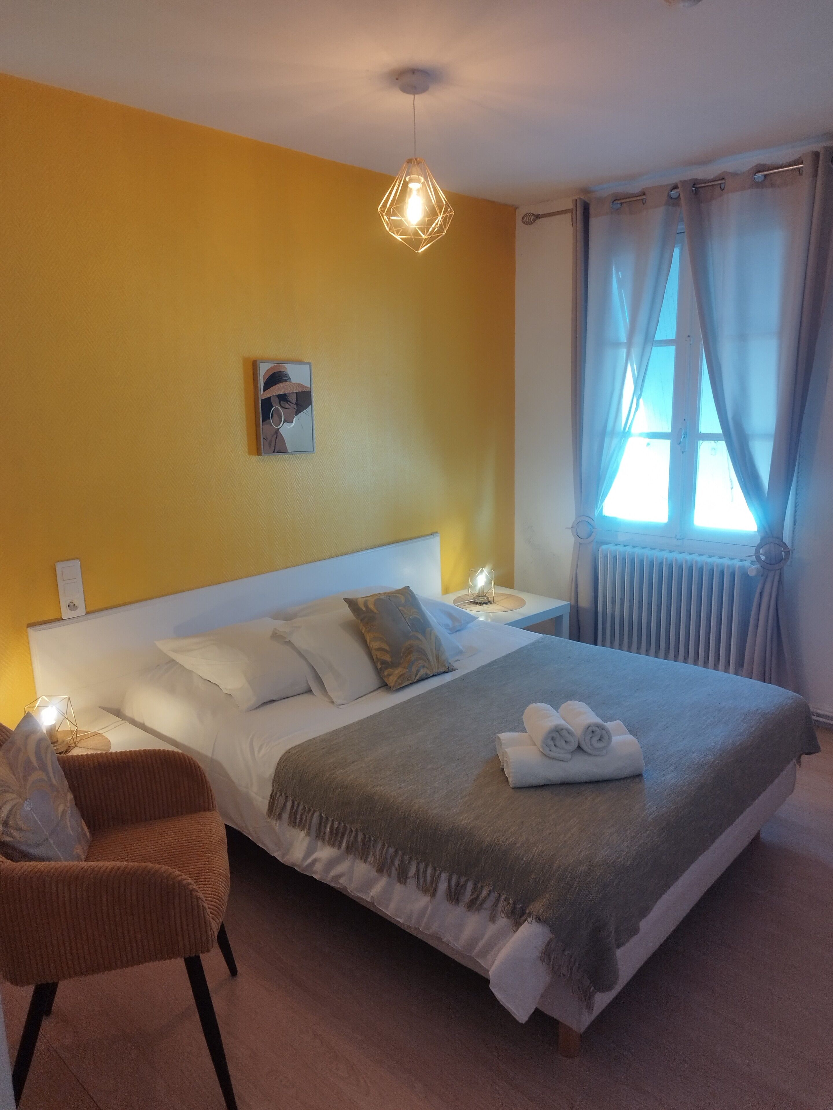 A hotel room with a large bed, a nightstand, a chair, a wall-mounted picture, and a window with curtains.