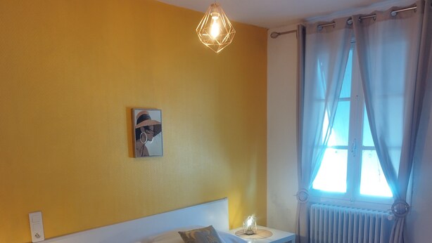 A hotel room with a large bed, a nightstand, a chair, a wall-mounted picture, and a window with curtains. - Hôtel les Thermes (Jonzac)