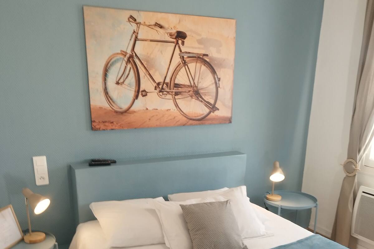 Cama bem arrumada com almofadas brancas e cinzas, um cobertor azul-esverdeado e toalhas dobradas sobre a cama. Há um quadro de uma bicicleta na parede acima da cama.