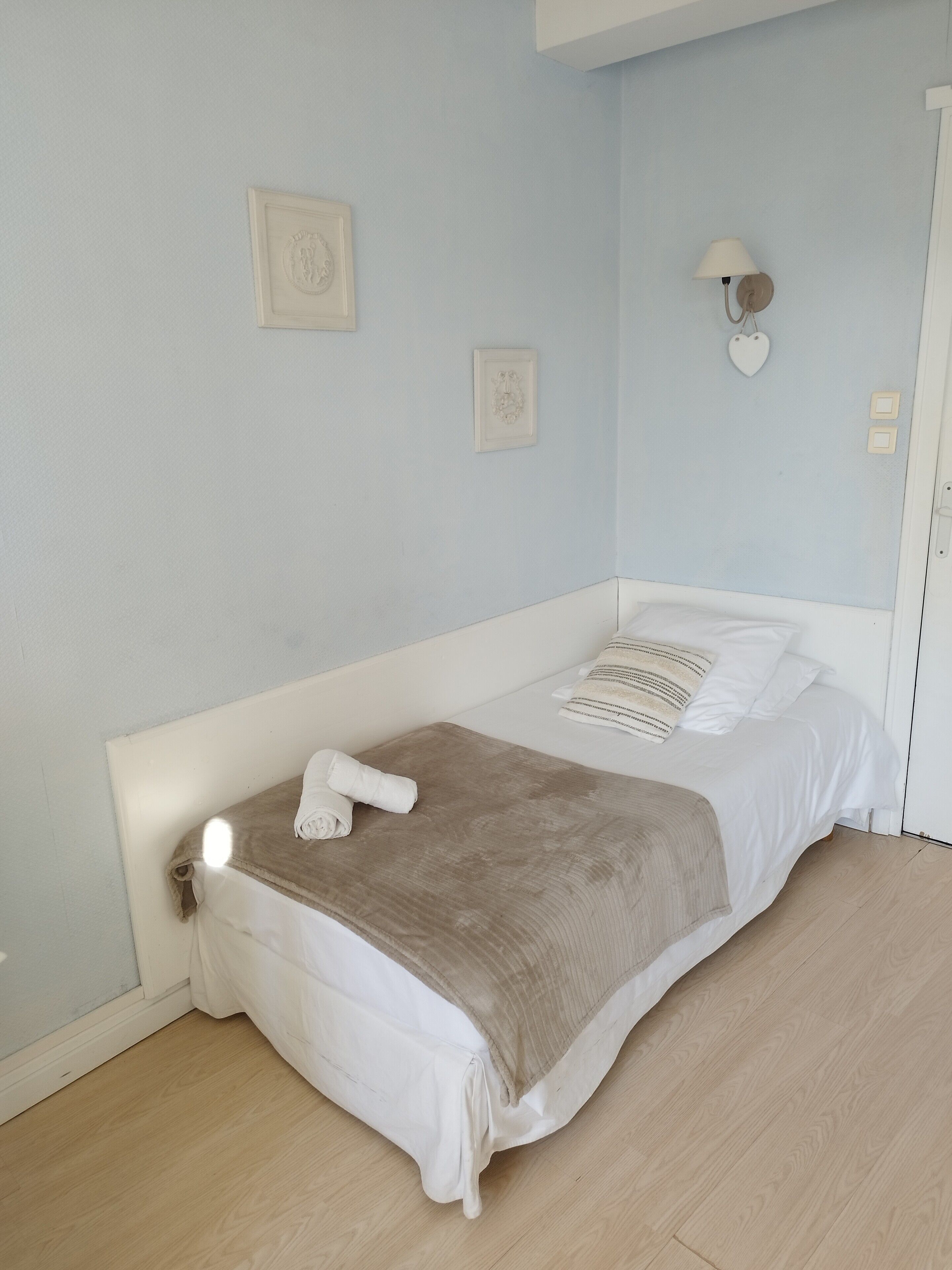 A single bed with a white headboard and footboard, a beige blanket, and a rolled towel on the bed. There are two wall-mounted light fixtures and two framed pictures on the wall.