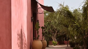 Uma trilha leva a um edifício com fachada rosa, grandes vasos de terracota e uma área externa arborizada com palmeiras.