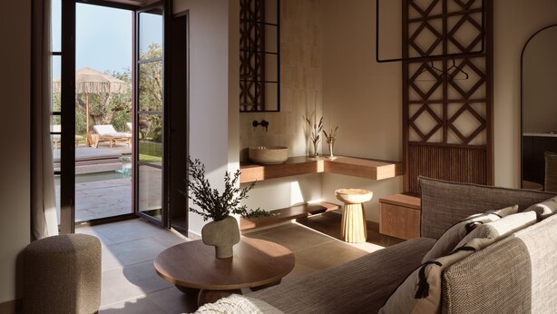 Uma sala de estar moderna com sofá, mesa de centro e vista para uma piscina e área externa com assentos através de uma porta de vidro.