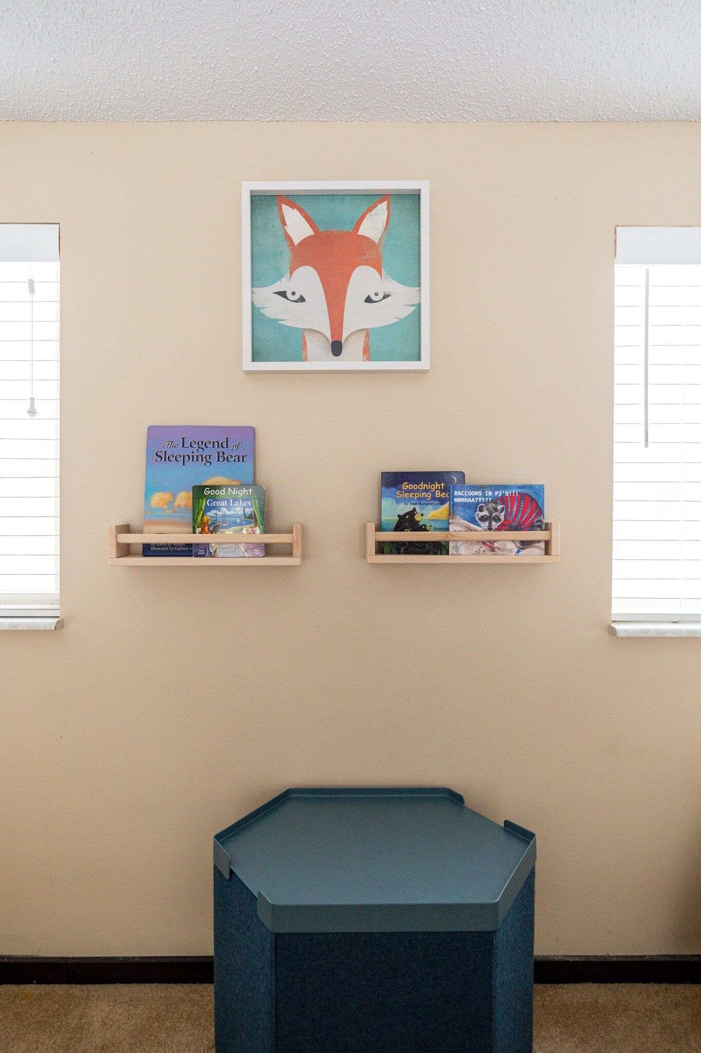 A room with a beige wall, a framed fox artwork, and two wooden shelves holding children's books.