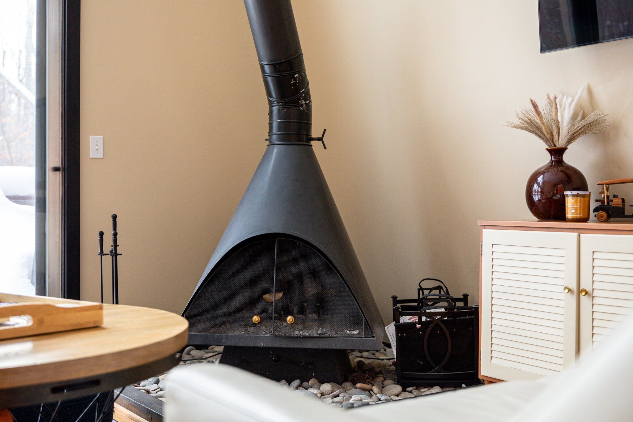 A room with a black wood-burning stove, a wooden table, a vase with dried plants, and a cabinet.