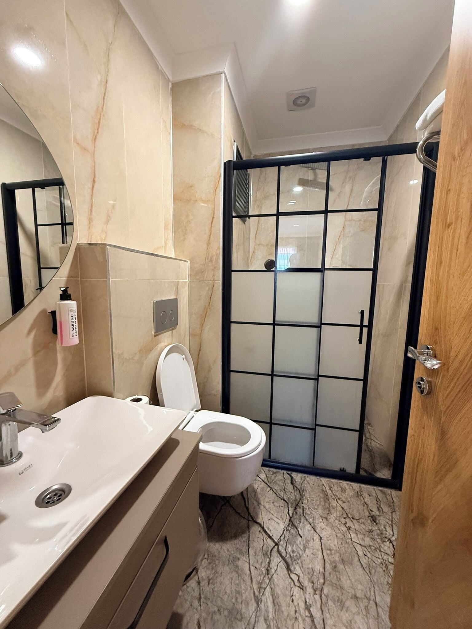 A bathroom with a marble floor, a white sink, a toilet, a large mirror, and a glass-enclosed shower.