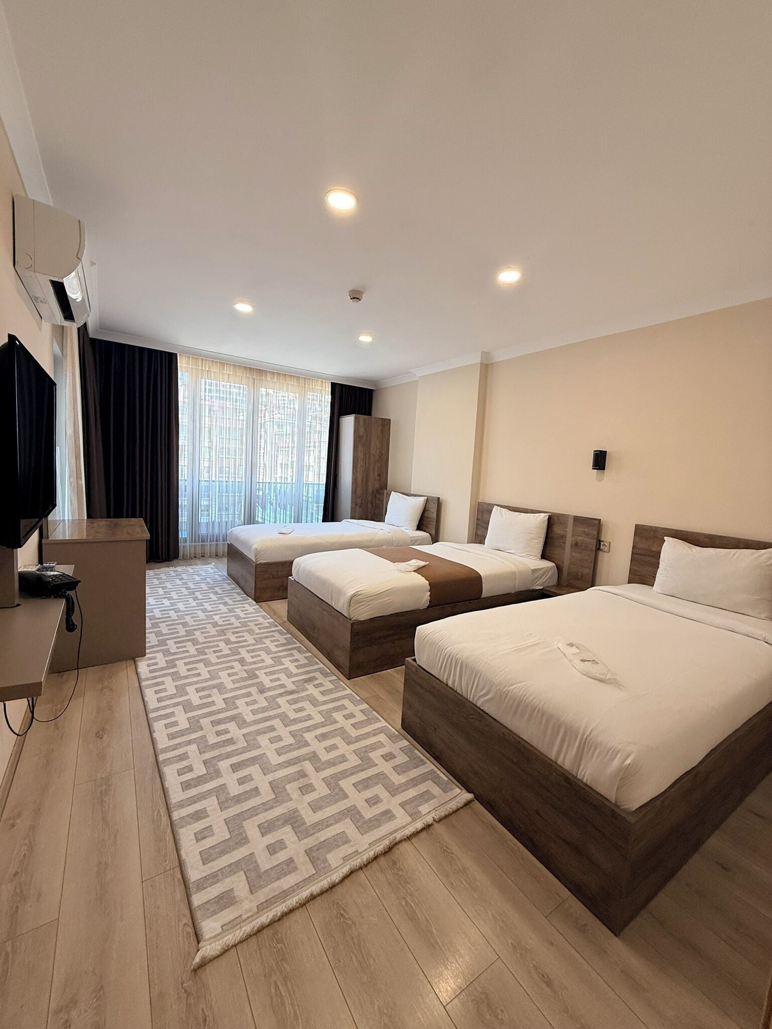 A hotel room with three beds, a desk, a TV, and a large window with curtains.