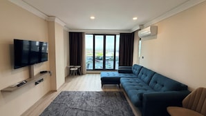 A modern living room with a flat-screen TV, a sectional sofa, a small coffee table, and a large window offering a view of the city. - Rena Grand Residence & Suite (Yomra)