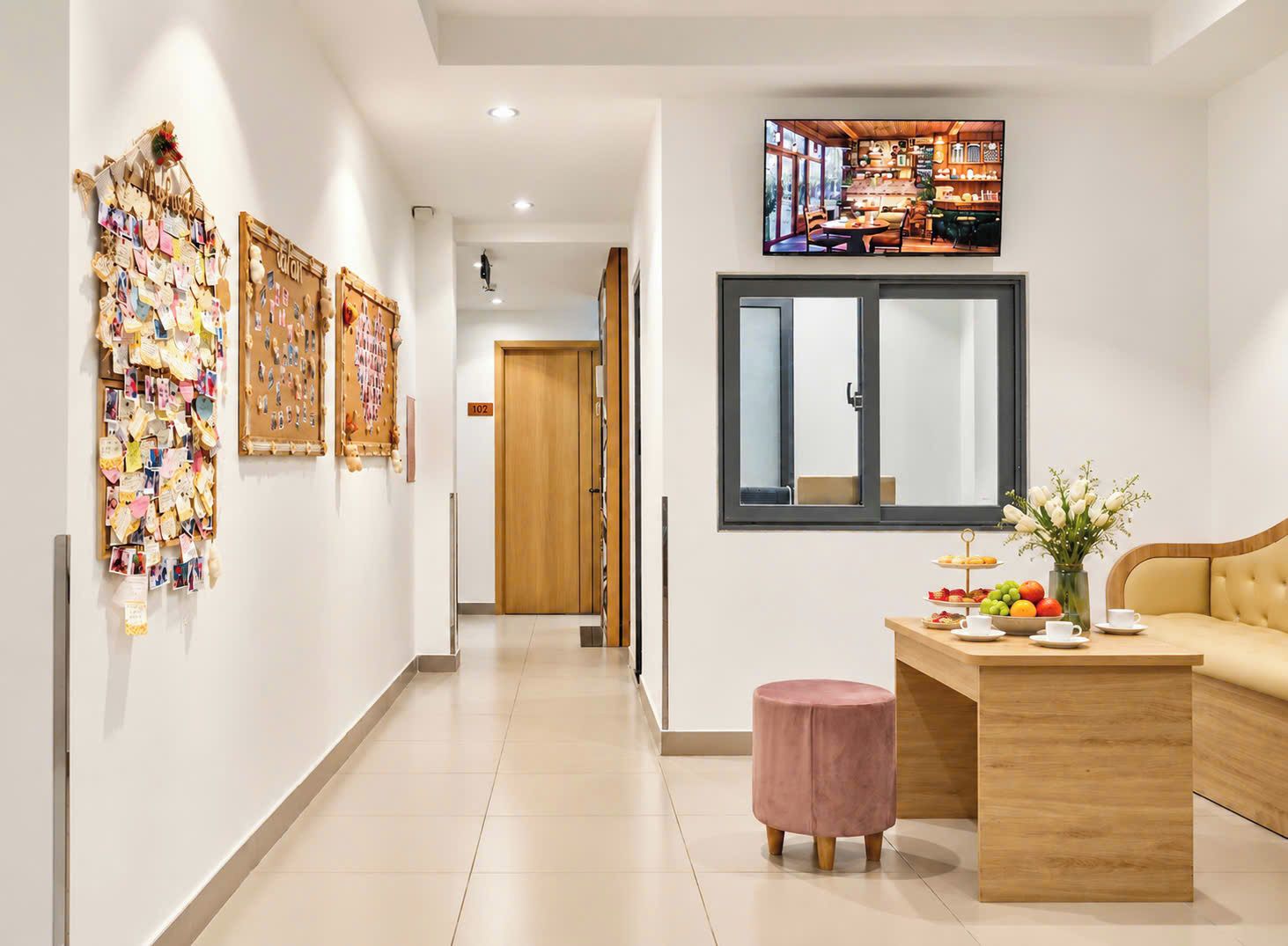A hotel corridor with a wooden reception desk, a pink ottoman, and a wall-mounted TV displaying a restaurant scene.