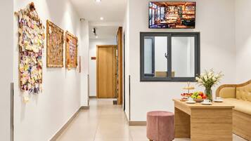 A hotel corridor with a wooden reception desk, a pink ottoman, and a wall-mounted TV displaying a restaurant scene.