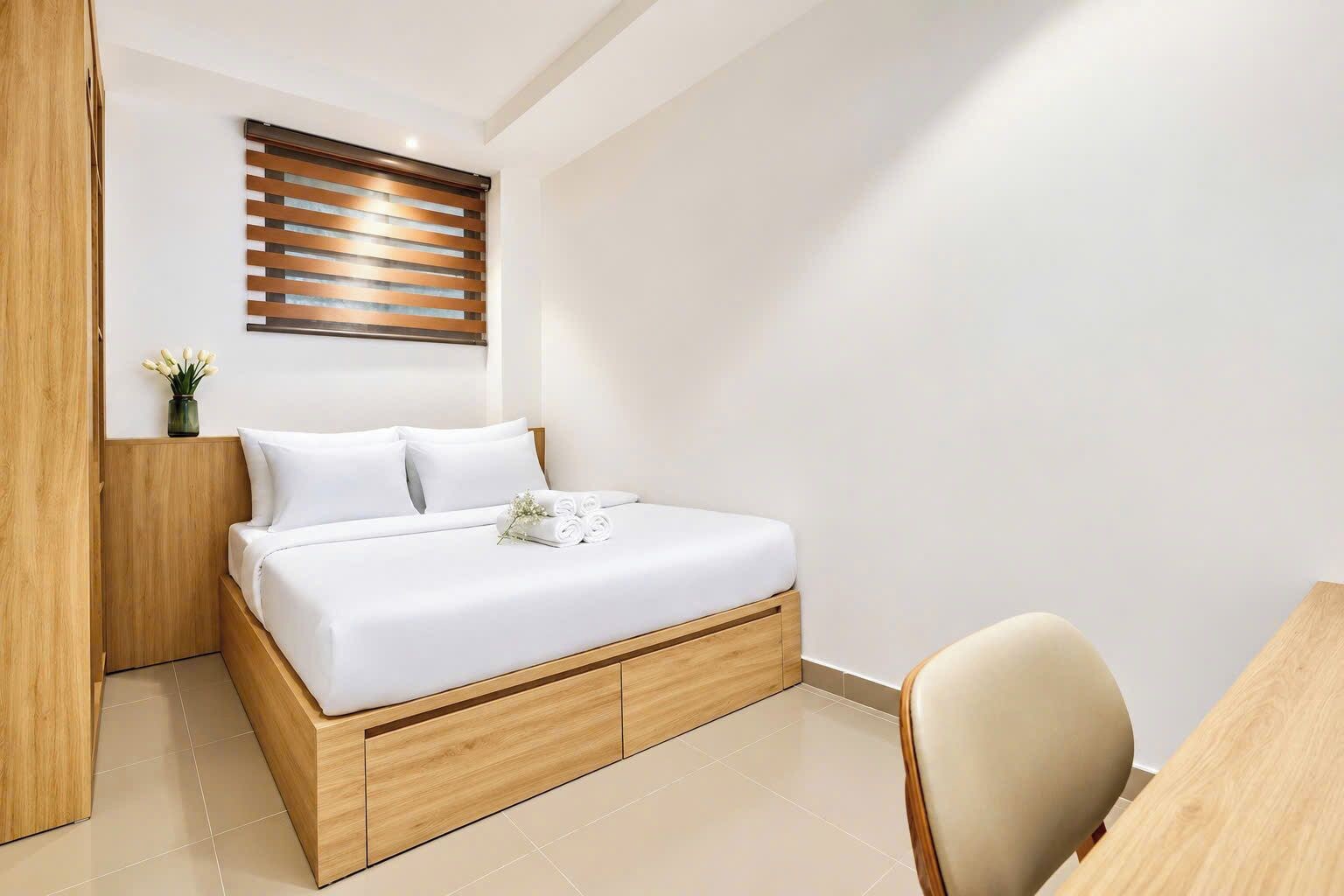 A modern bedroom with a wooden bed frame, white bedding, a wooden headboard, a bedside table with a vase of flowers, and a chair.