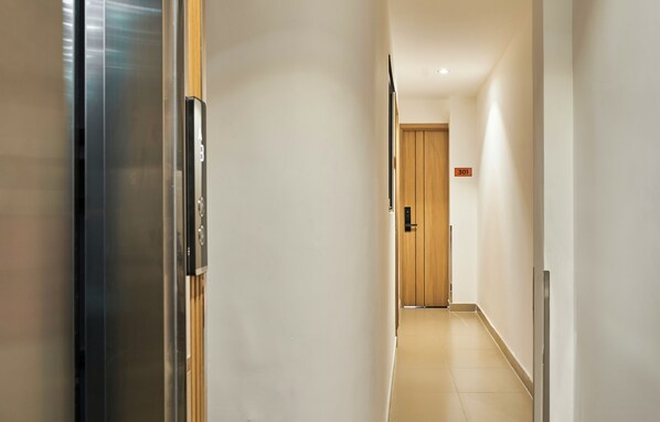 A modern hotel hallway with a wooden door at the end, a metal elevator, and a sign indicating the direction to the room. - Mihn Suites Ben Thanh (Ho Chi Minh City)