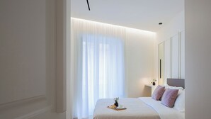 Un dormitorio moderno con una cama grande, una mesita de noche con lámpara y cortinas translúcidas.