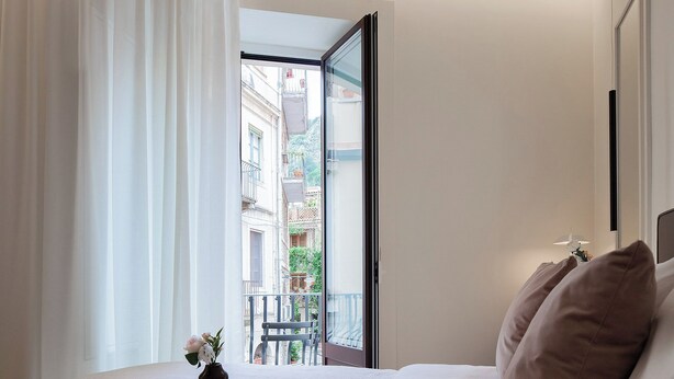 Habitación de hotel con una cama, una bandeja con croissants y una taza, y vistas a edificios desde una puerta abierta.