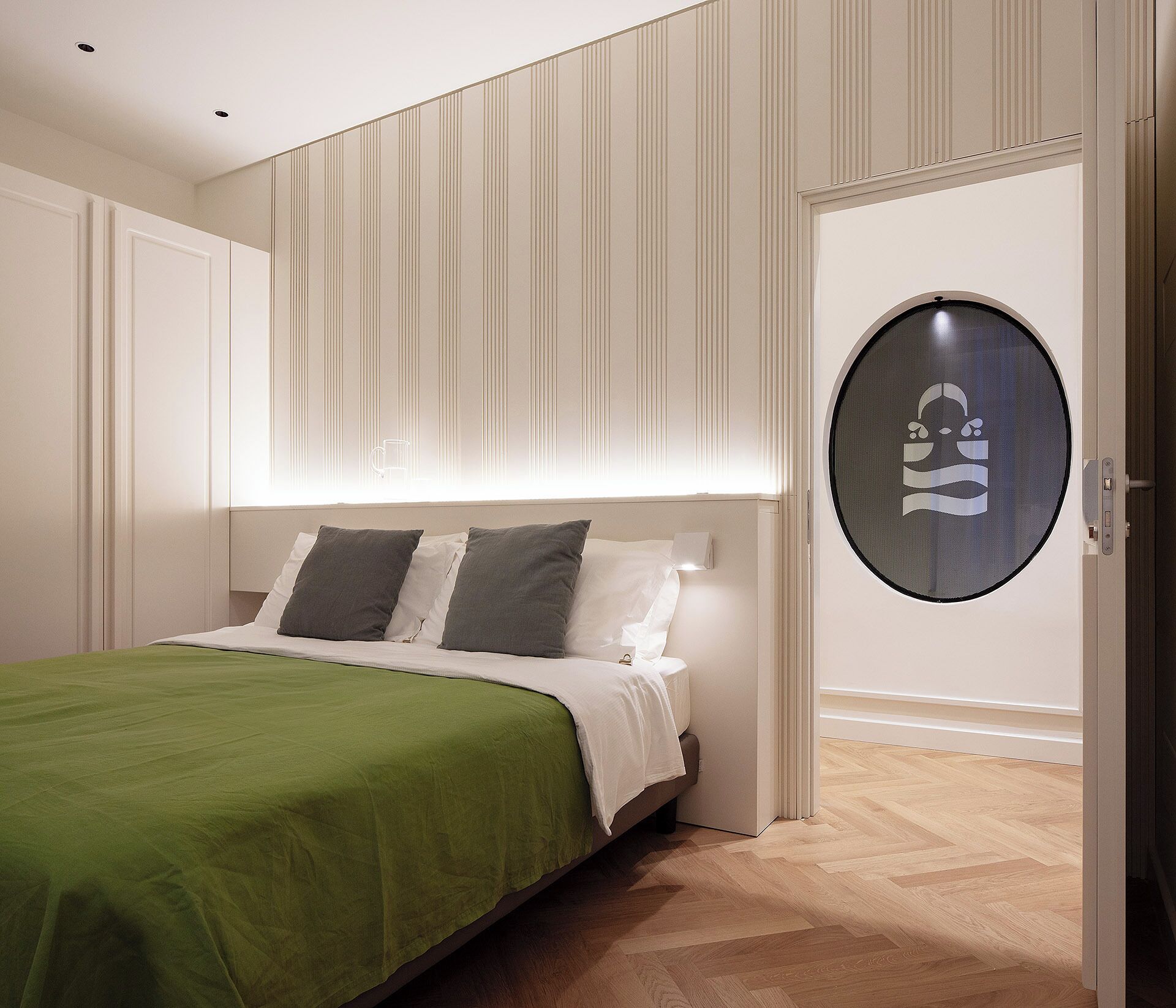Un dormitorio con una cama, una colcha verde, un cabecero blanco y un espejo redondo en la pared.