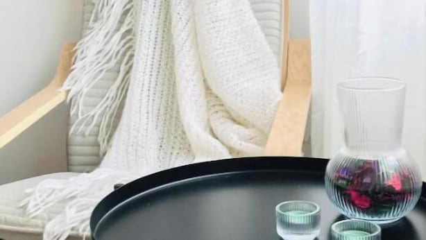 Una silla con un cubrecolchón blanco de punto, una mesa negra con un florero de vidrio y dos vasos de vidrio, y un piso de madera.