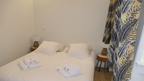A neatly made bed with white linens, a bedside table with a lamp, and a tropical leaf-patterned curtain.