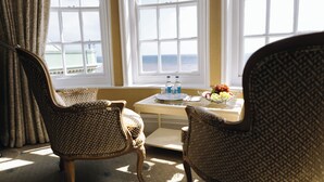 Una habitación con sillas de mimbre, una mesita con una bandeja de frutas y botellas, y vista al mar a través de amplias ventanas.