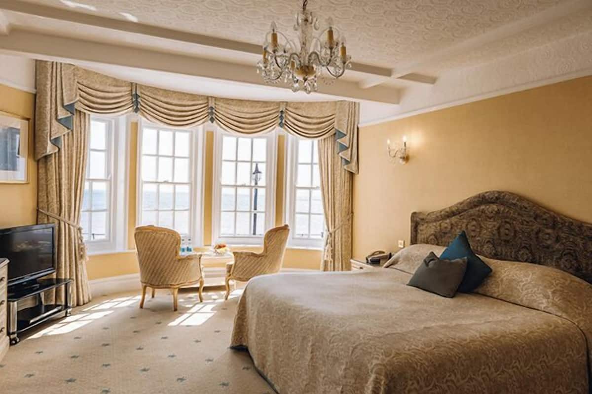 A hotel room with a large bed, a television, a desk with chairs, and a chandelier.