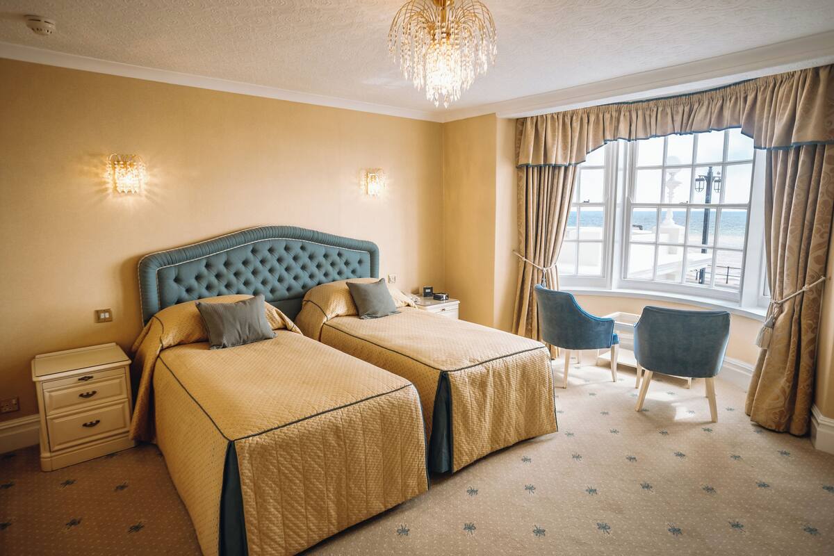 A hotel room with two beds, a chandelier, a window with curtains, a chair, and a nightstand.