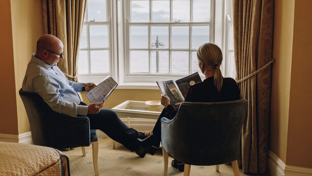Dos personas sentadas en sillones junto a una ventana con vista al mar.