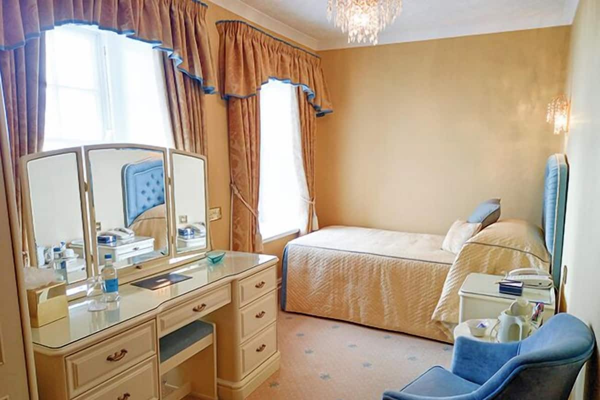 A hotel room with a bed, a desk with a mirror, a chair, and a bedside table.