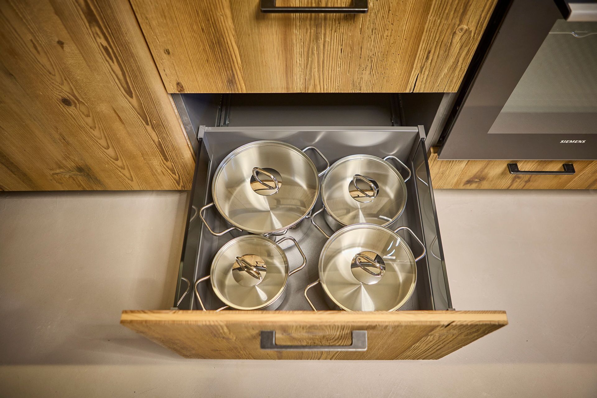 Eine Küche mit vier Edelstahl-Töpfen auf einem Herd, einem Holzschrank darüber und einem Siemens-Backofen zur Rechten.